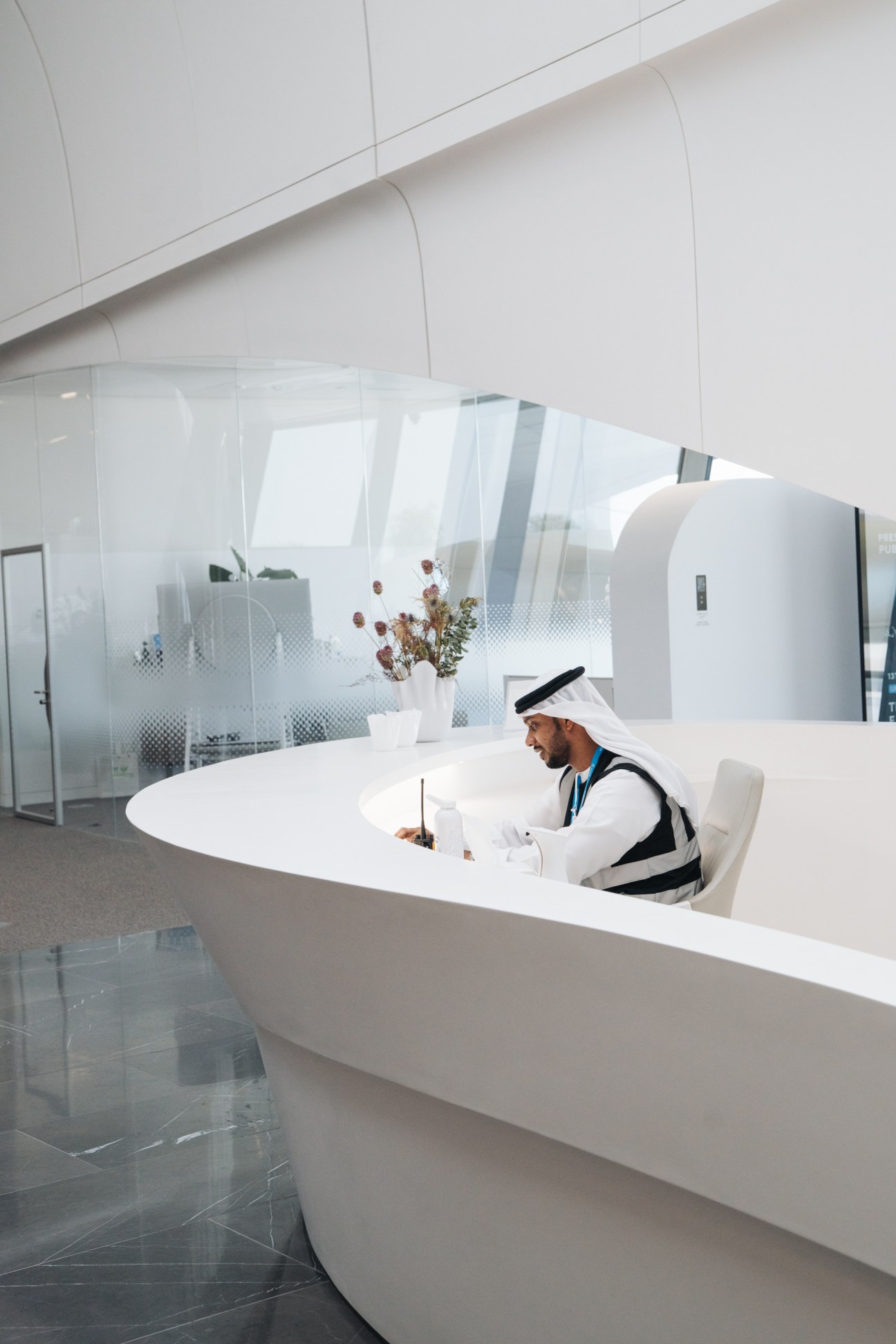 A man in traditional Emirati dress works at a curved white reception desk in a minimalist interior with frosted glass partitions.