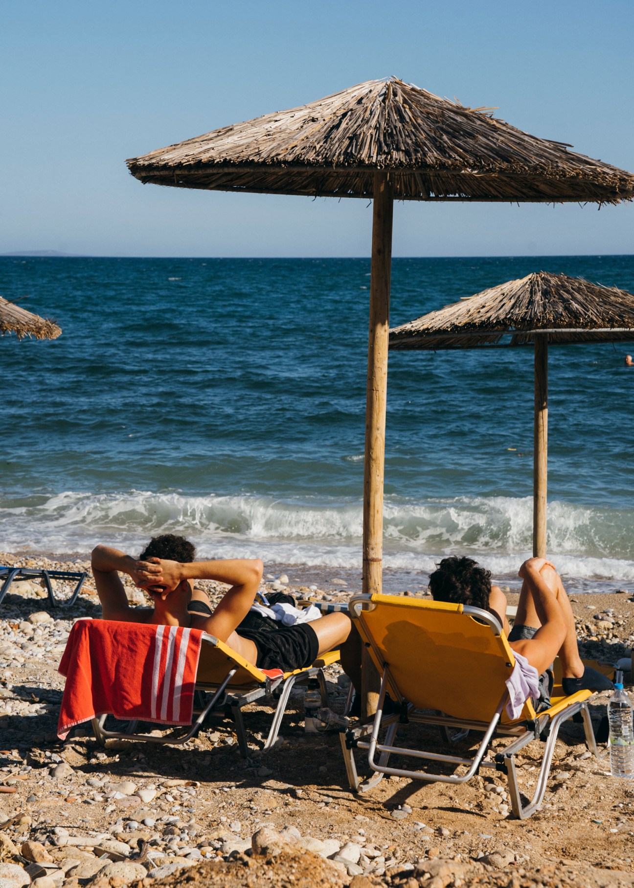 Two people sunbathing on lounge chairs under thatched umbrellas at a pebble beach with waves in background