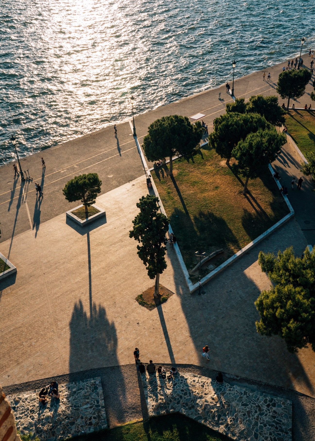 Aerial view of Thessaloniki's waterfront promenade with beach, green spaces, scattered trees, and people strolling along the Aegean Sea.