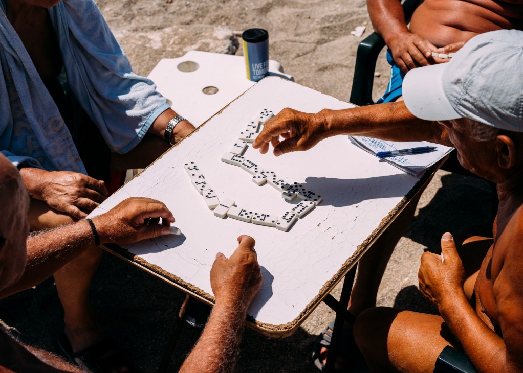 People playing dominoes around a small table outdoors in sunny weather