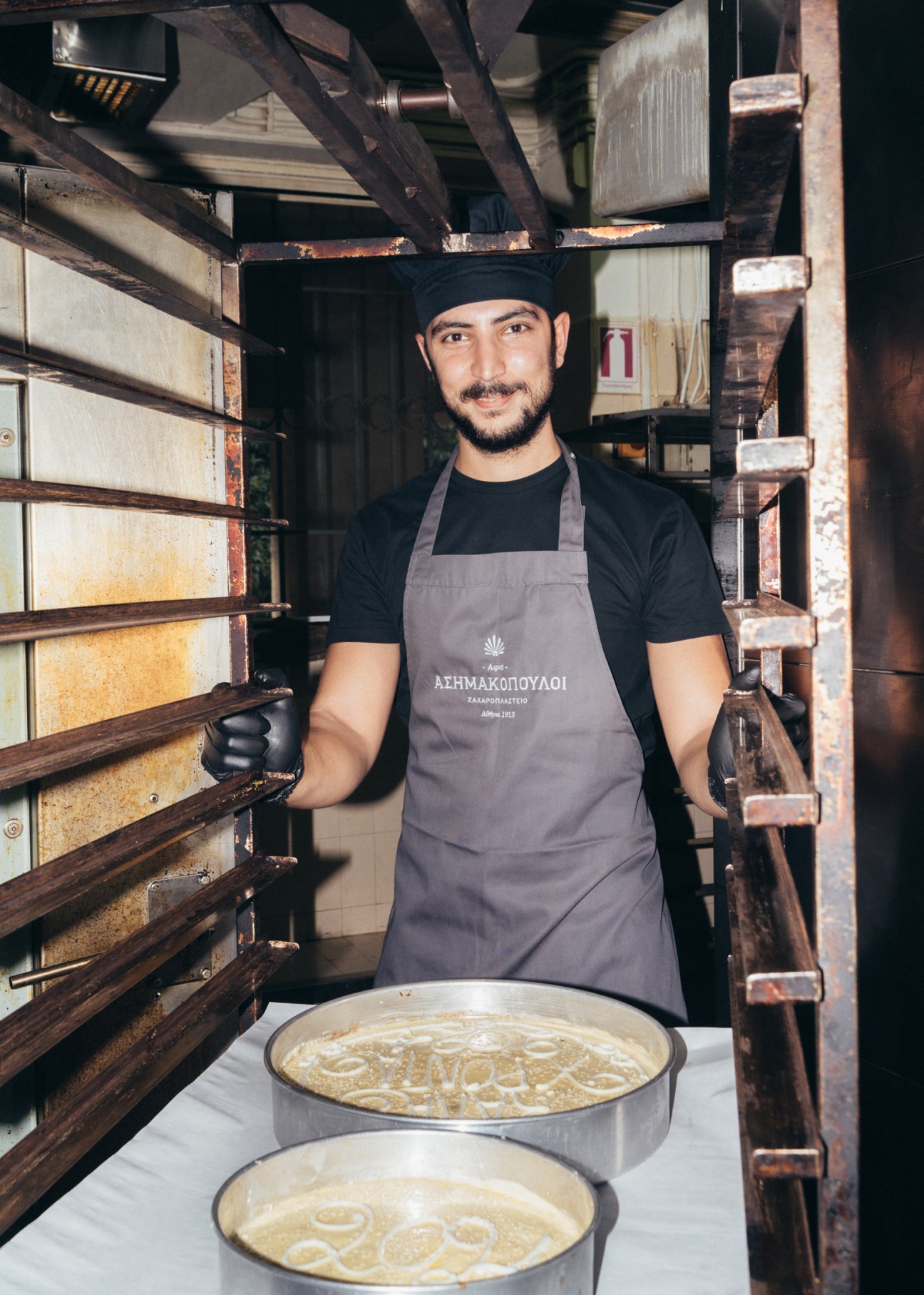 Baker at Asimakopouloi in Athens holds vasilopita cakes by industrial ovens.