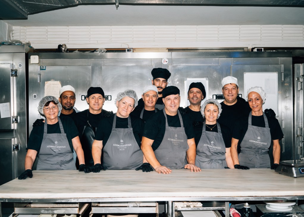 Team of bakers wearing hairnets and aprons standing together in Asimakopouloi bakery kitchen in Athens.