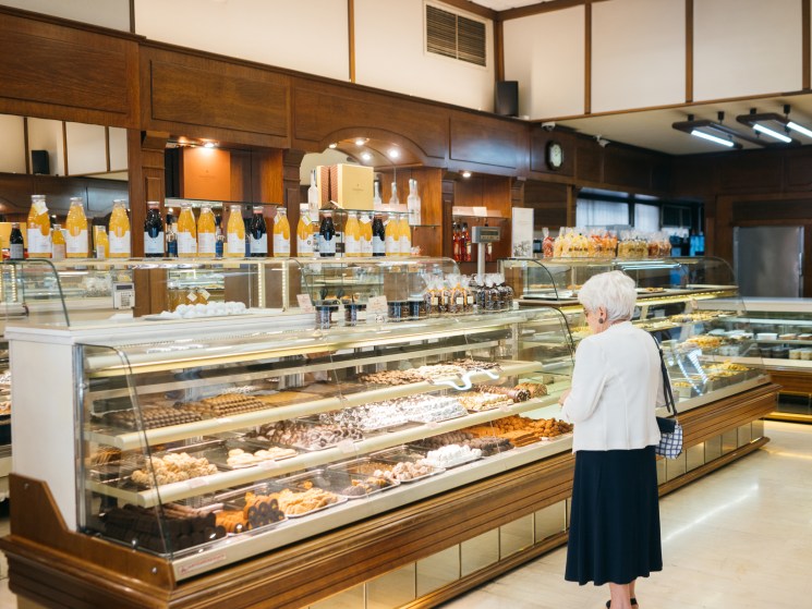 Elderly woman in white blouse browsing display cases filled with pastries and chocolates in upscale bakery