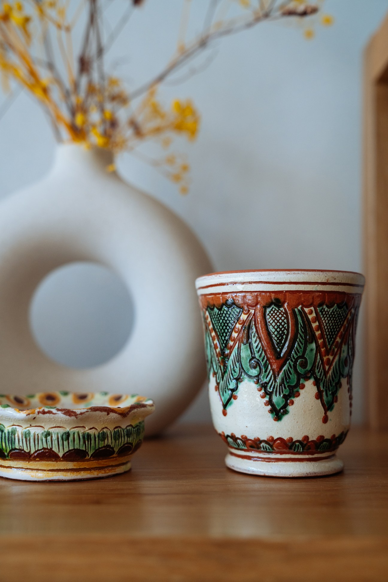 Hand-painted ceramic cup with red and green geometric patterns next to a white vase with dried yellow flowers.