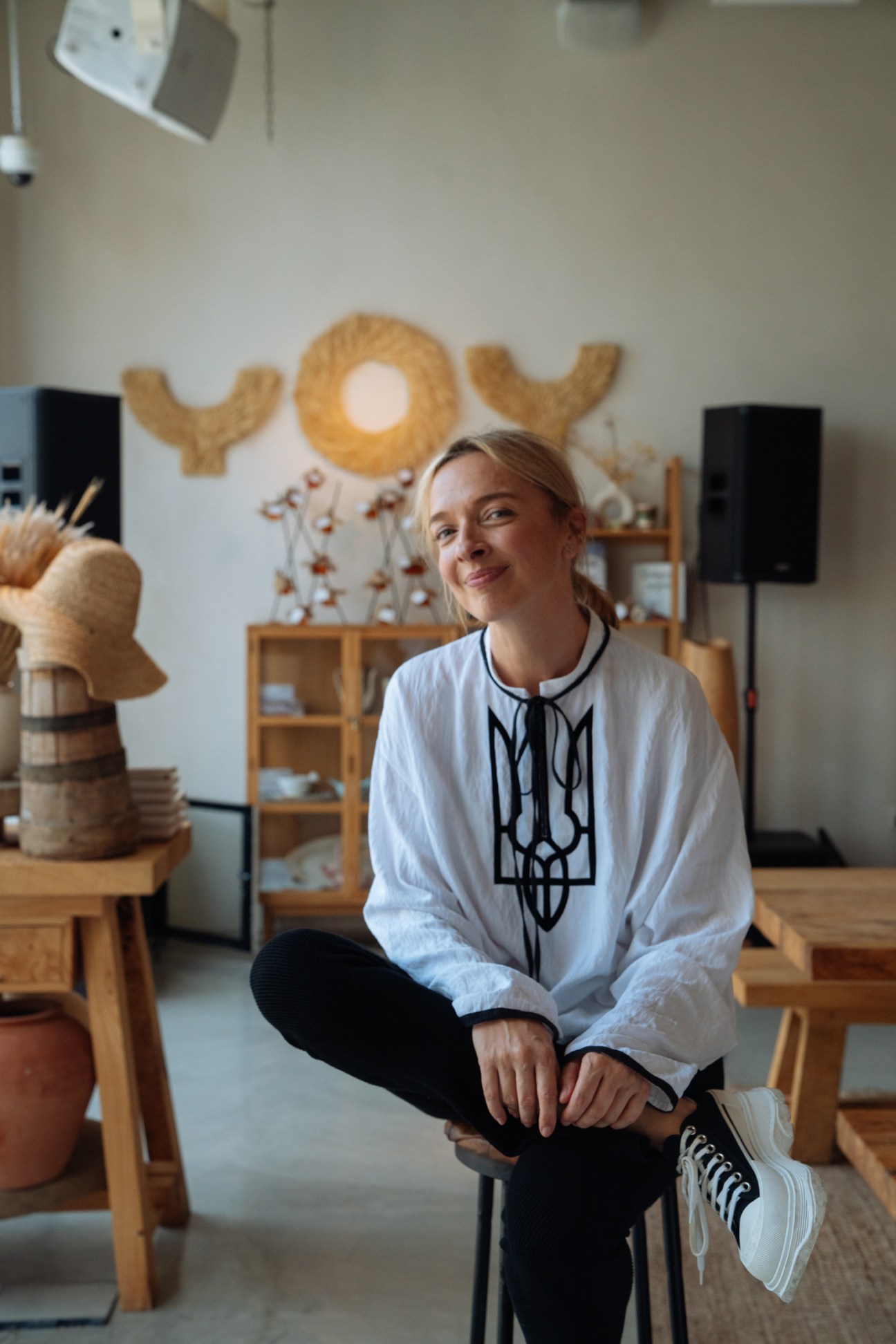 Woman in white shirt with black embroidered neckline sitting in modern furniture showroom with wooden shelving and woven wall art.