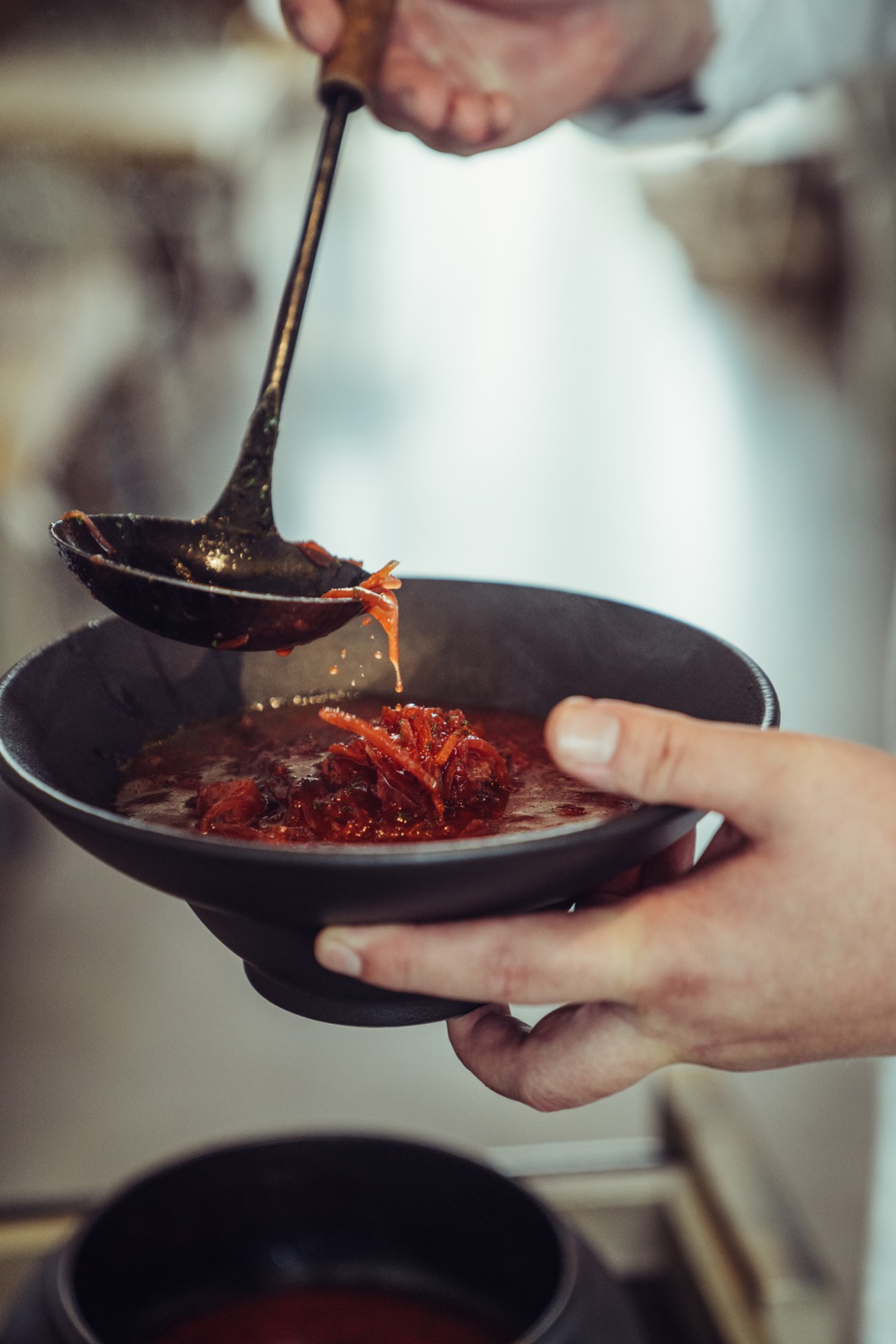 Hands holding a black bowl while ladling beetroot borscht soup with a spoon.