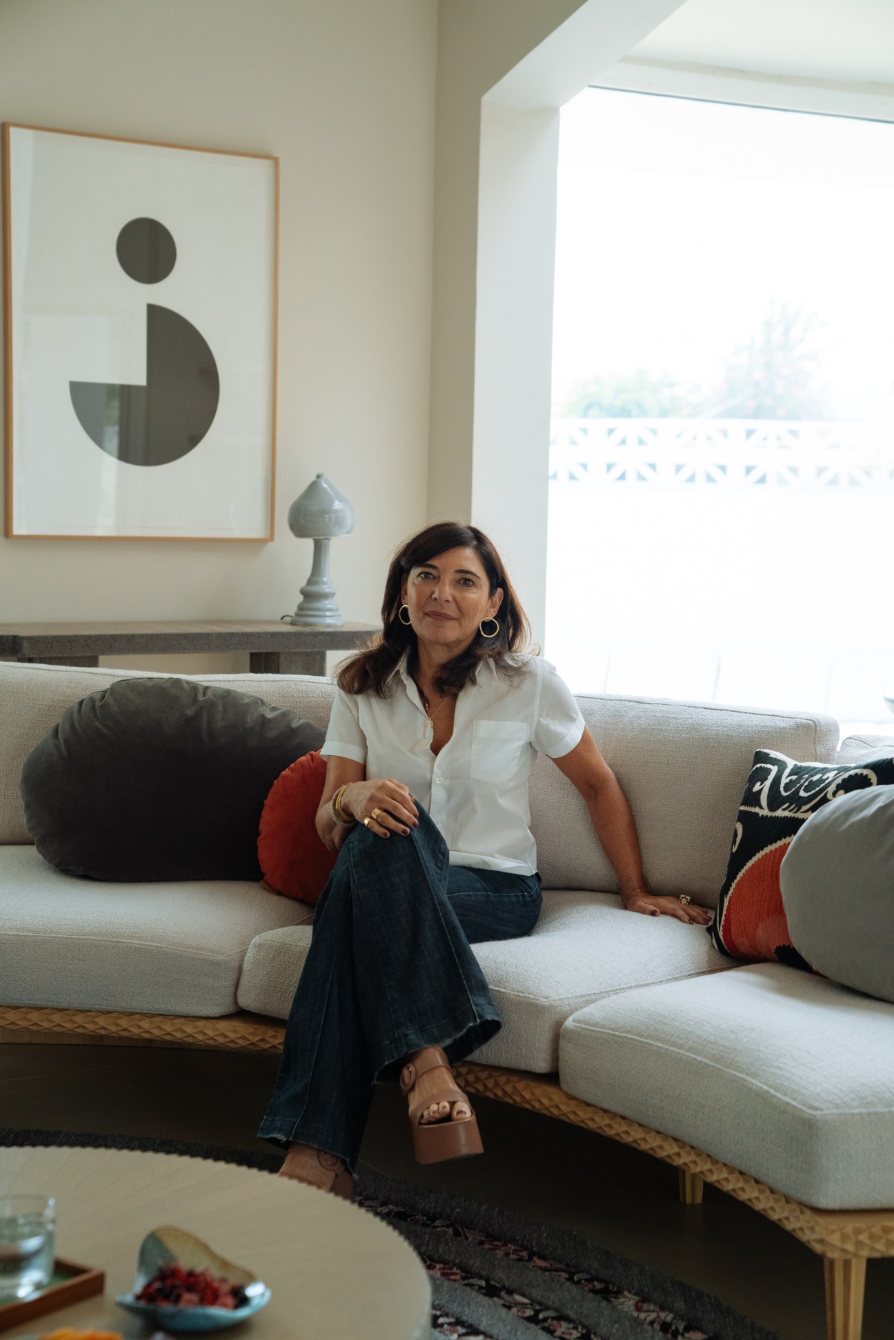 Lebanese designer Nada Debs sits on a cream sofa in her Dubai home, surrounded by minimalist furnishings and geometric artwork.