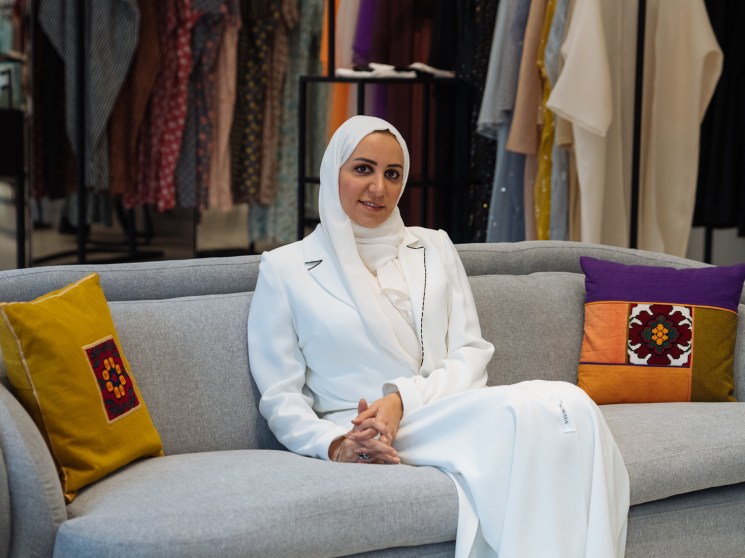Hala Al Gergawi, founder of Tea Before Noon, sits on a grey sofa in front of clothing racks displaying colorful abayas and garments.