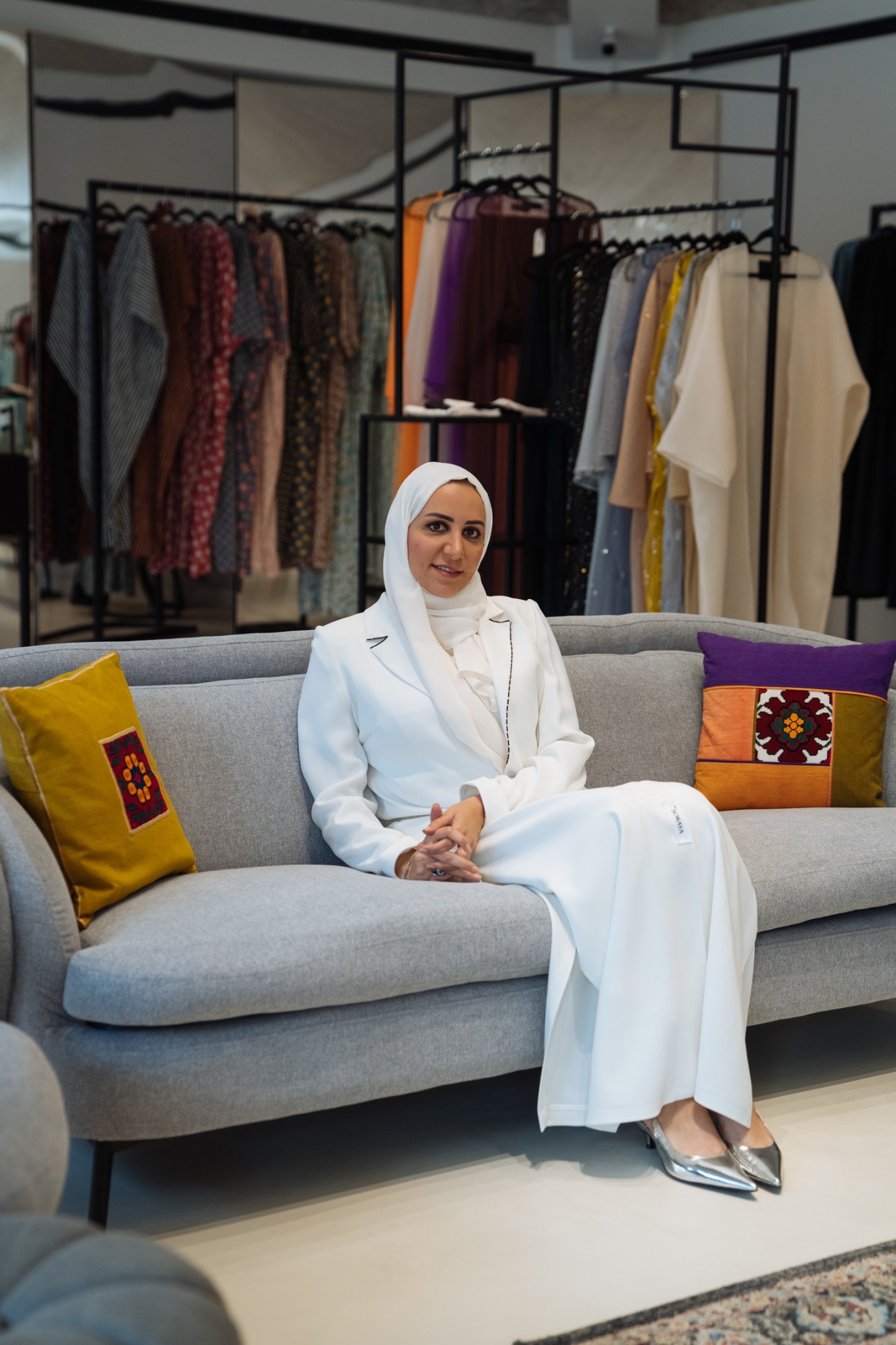 Hala Al Gergawi, founder of Tea Before Noon, sits on a grey sofa in front of clothing racks displaying colorful abayas and garments.