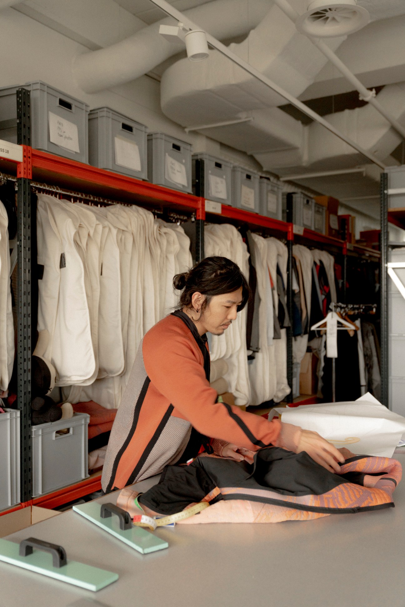 Person in orange shirt examining textiles at workspace with shelving of garments and storage boxes