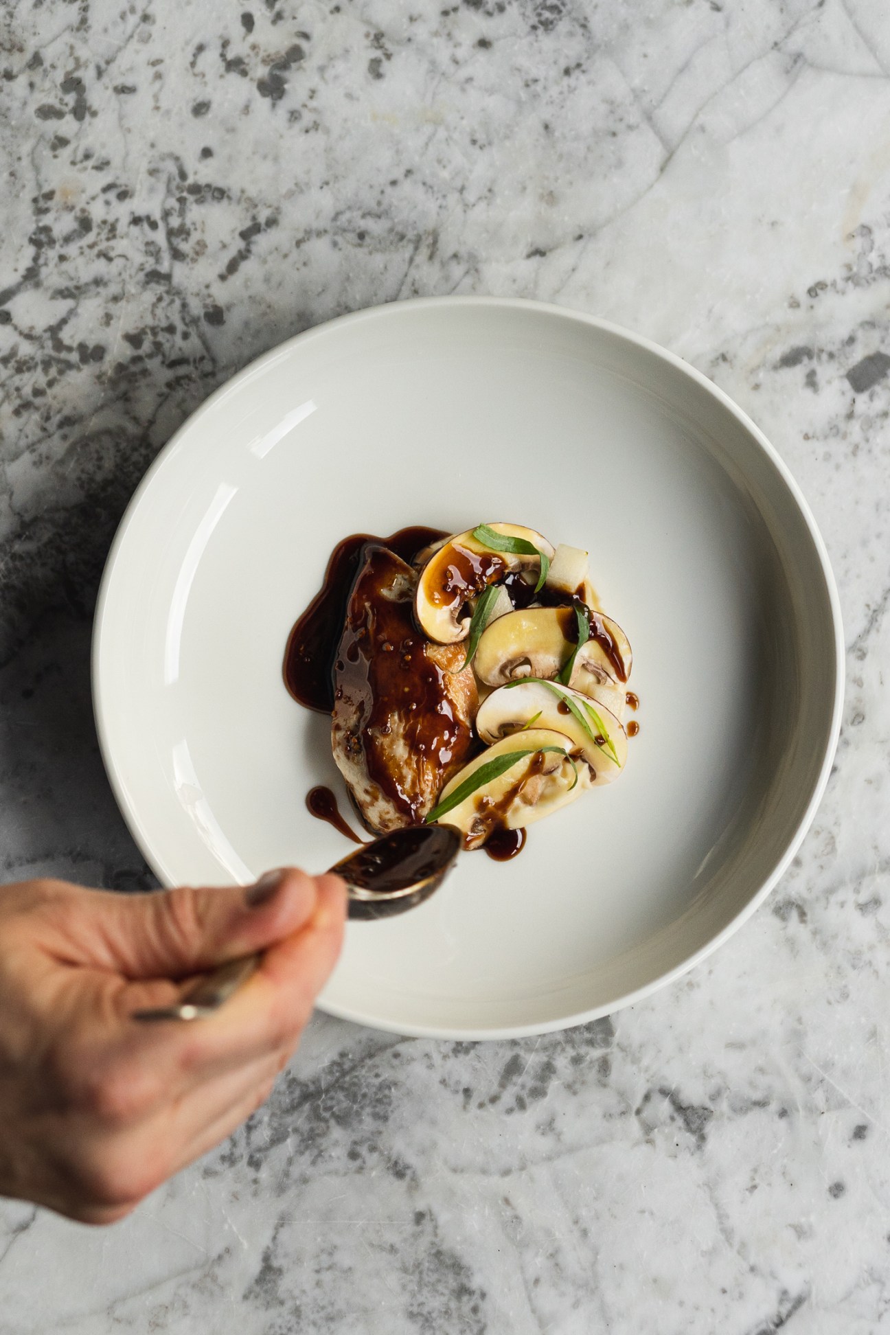 Plated dish with seared meat, mushrooms, apple slices and dark sauce on white ceramic plate held over marble counter.