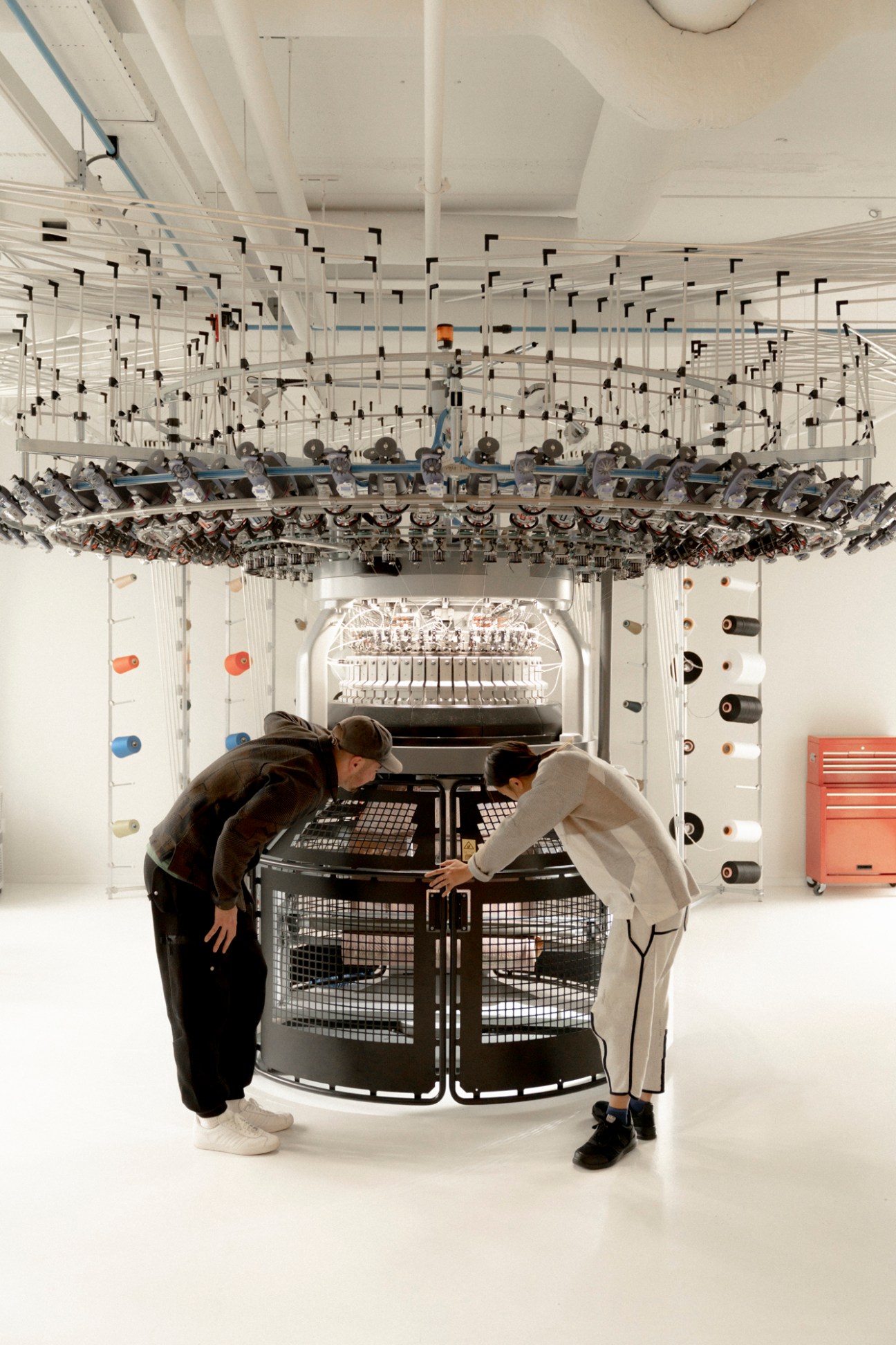 Two people examining an industrial knitting machine in a white studio space with complex textile machinery overhead