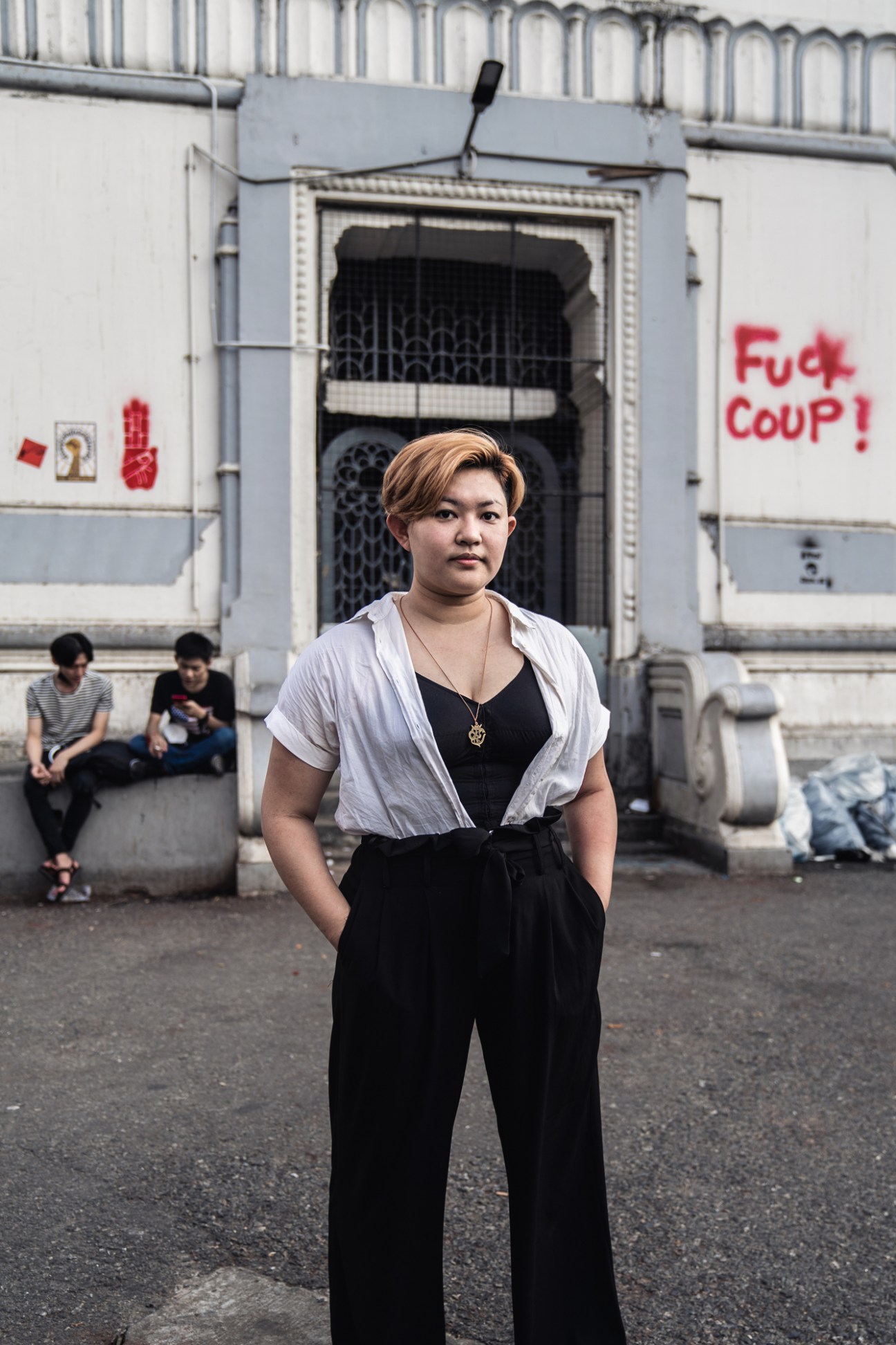 Aye Min Thant standing on a street in front of a building with graffiti reading 'Fuck coup!'