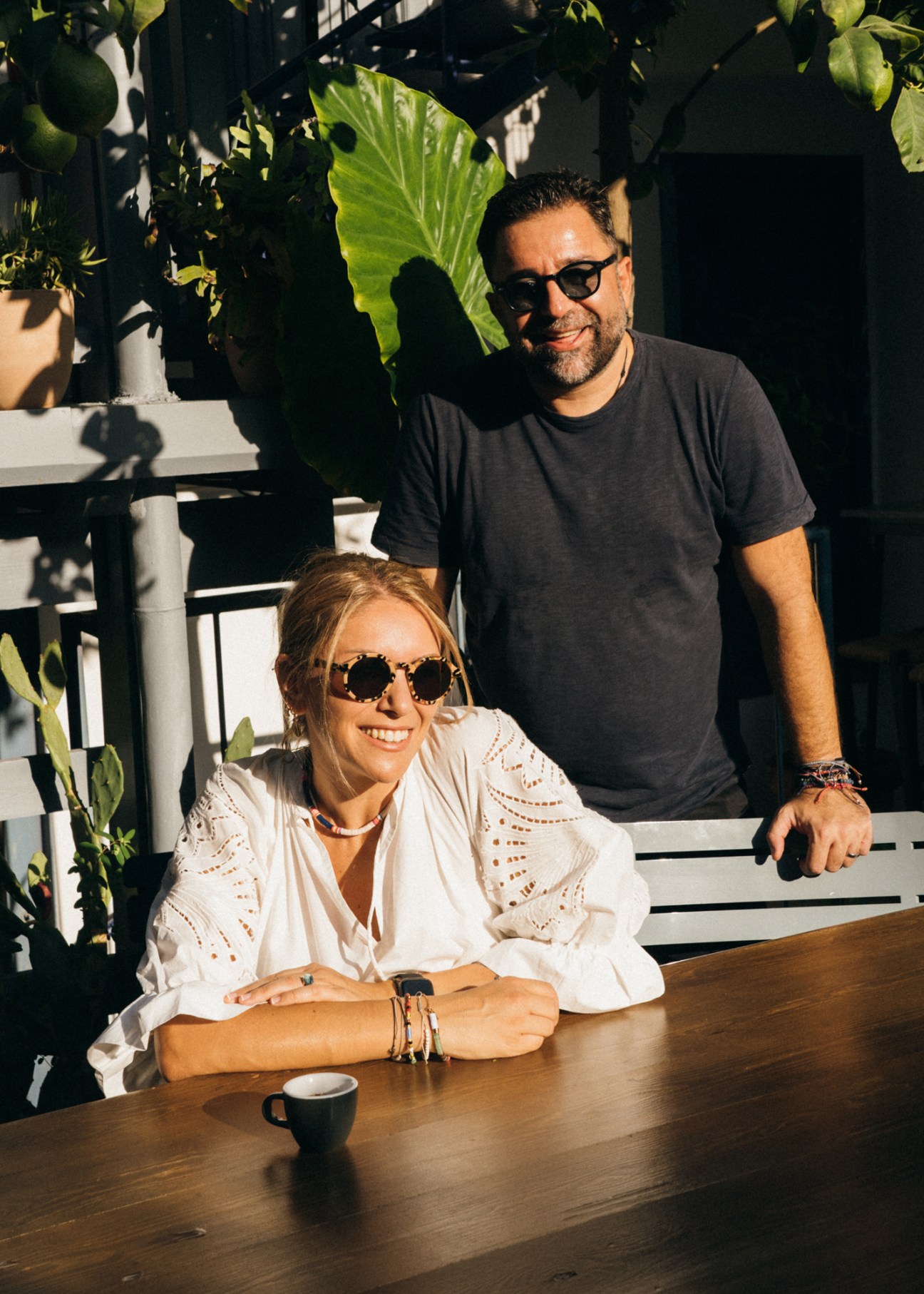 Woman in white eyelet top and man in gray t-shirt smiling at outdoor table with coffee cup