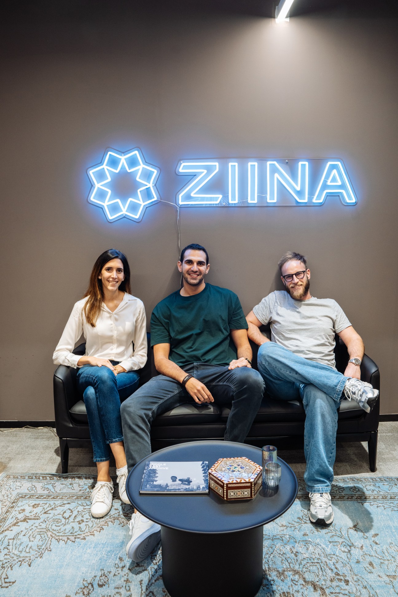 Three people sit on a black sofa beneath a blue neon Ziina logo sign.