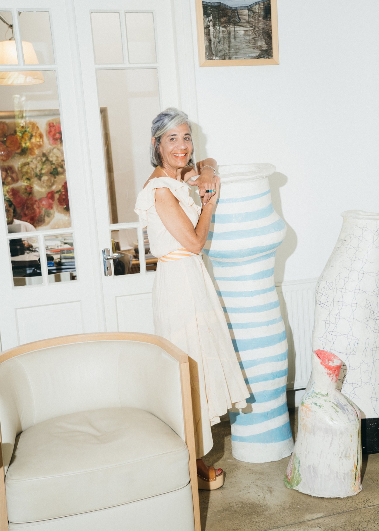 Woman with gray hair wearing white top standing in bright whitewashed studio with striped ceramic vessel