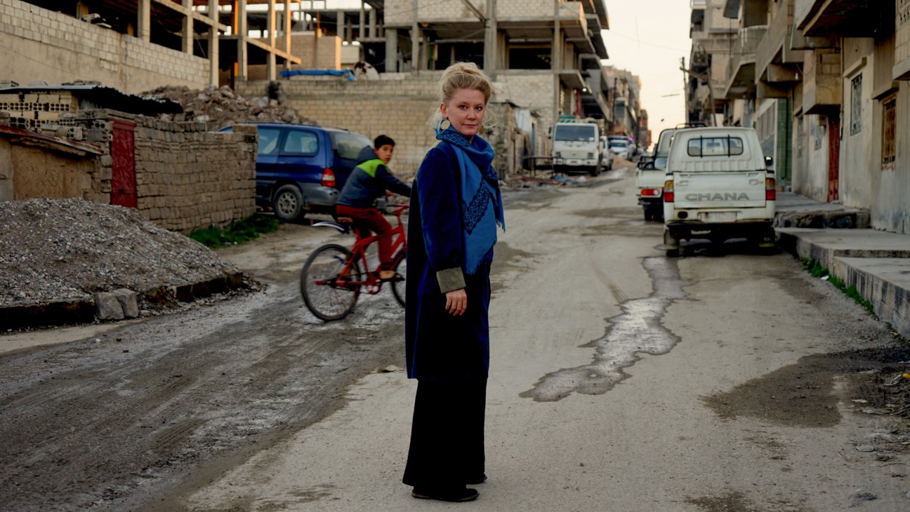 Puk Damsgård standing on a damaged street with concrete buildings and vehicles in the background