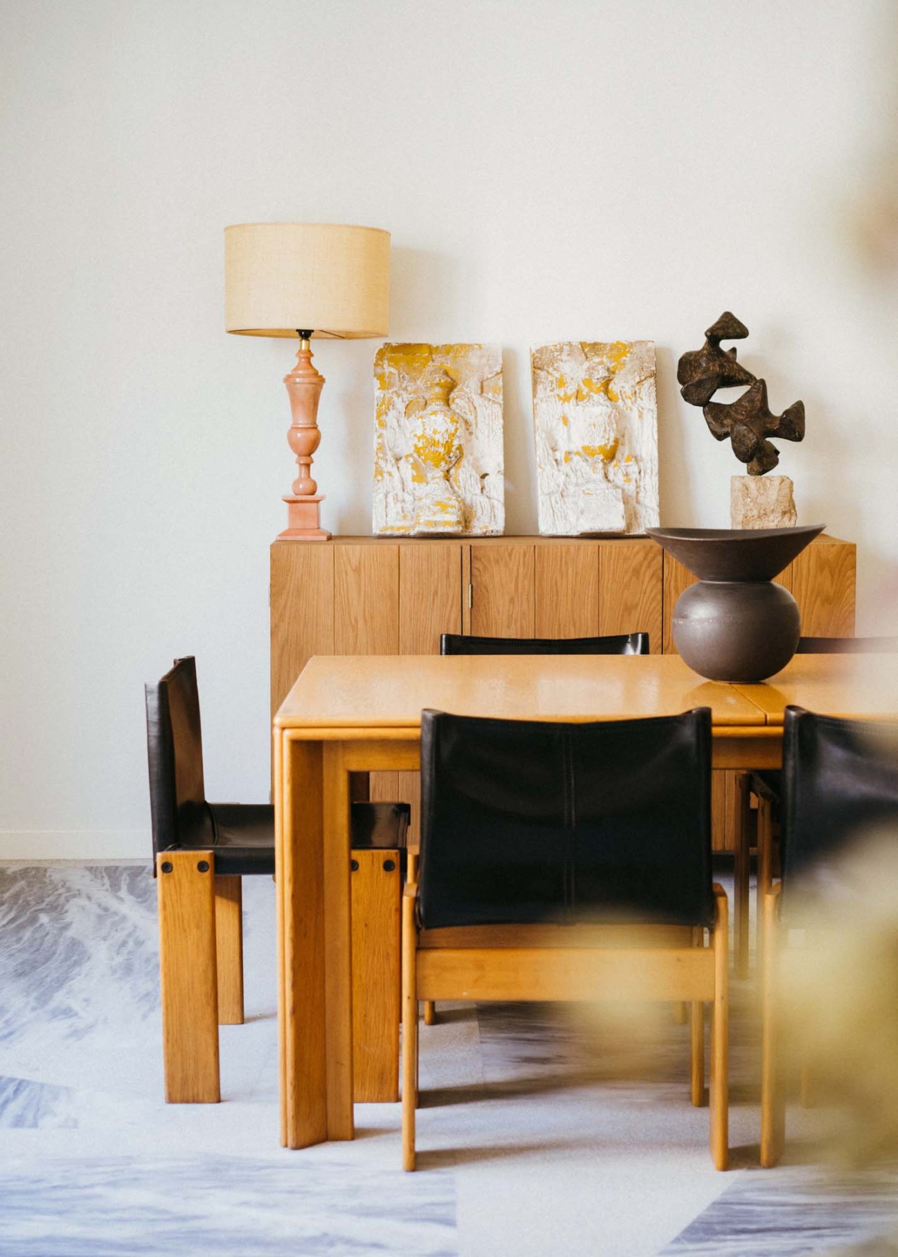 Dining area with wooden table and black chairs by Afra and Tobia Scarpa, styled with a lamp, artwork, and decorative objects on a marble floor.