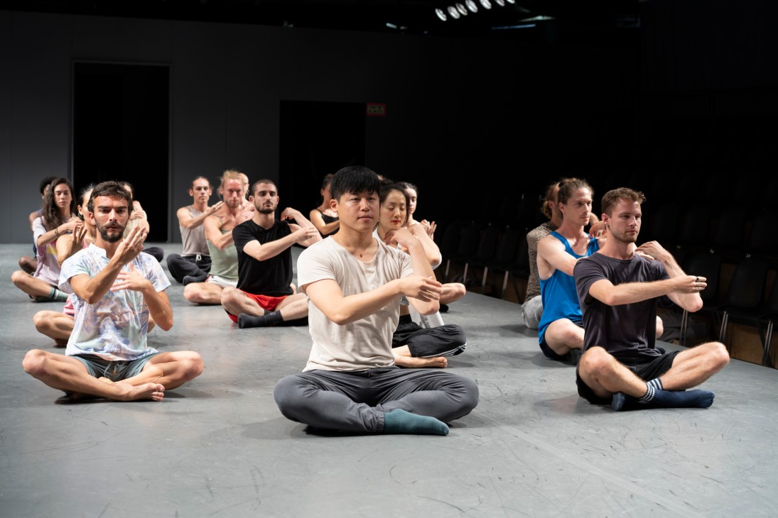 Dancers sitting cross-legged on studio floor performing synchronized movements during rehearsal
