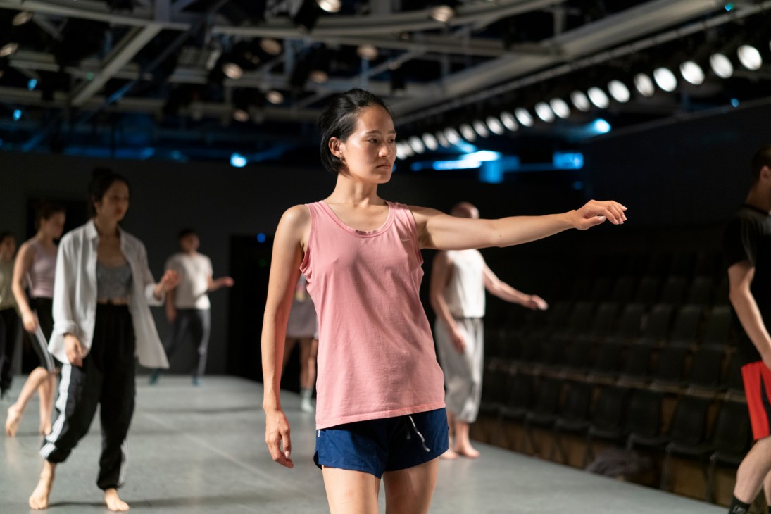 Dancer in pink tank top extends arm during rehearsal in modern studio with other dancers in background