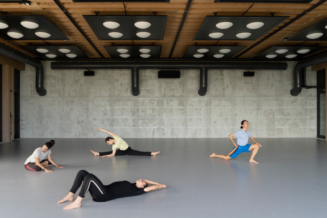 Four dancers stretching and warming up on the grey floor of a modern dance studio with industrial ceiling