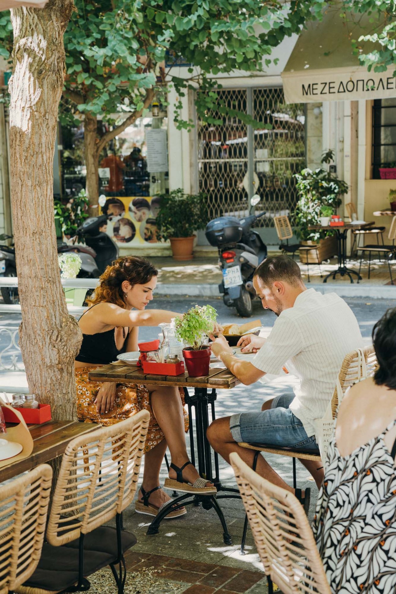 Two people dining outdoors at a table on a tree-lined street in Athens' Kypseli neighborhood.