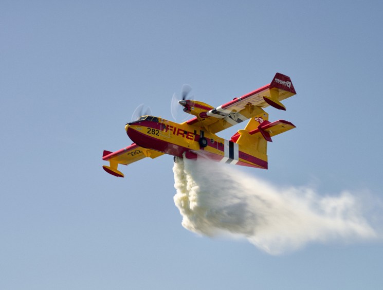 Yellow and red firefighting aircraft dropping water or retardant while flying against a blue sky