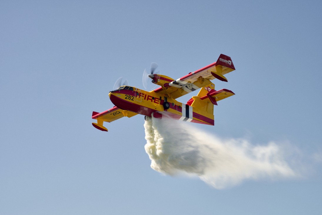 Yellow and red firefighting aircraft dropping water or retardant while flying against a blue sky