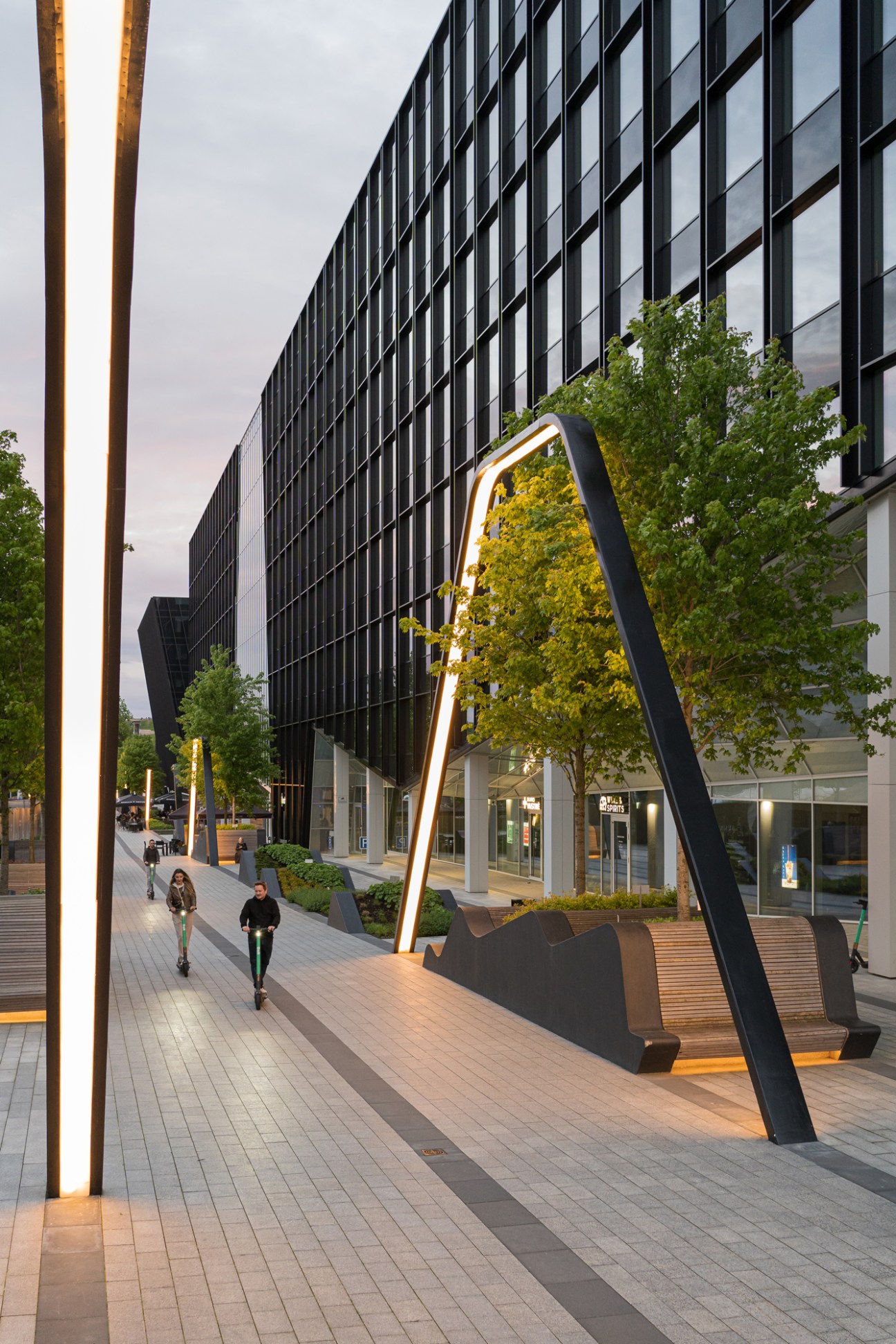 Vilnius Plaza features curved black metal structures with golden lighting and wooden seating beneath trees alongside a modern building.