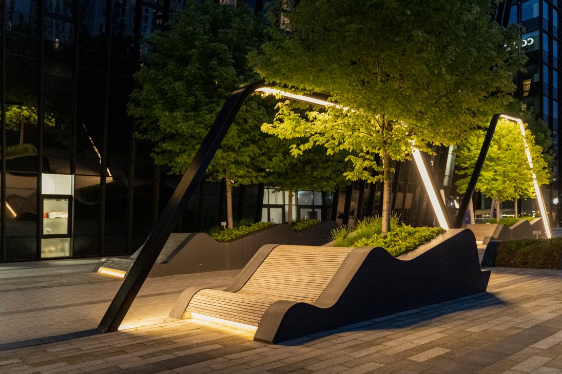 Illuminated curved wooden seating structures with overhead lighting in Vilnius Plaza at night, surrounded by trees and buildings.