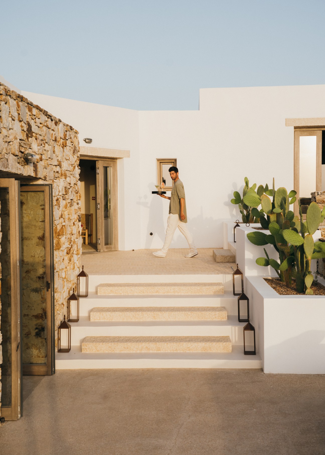 A man in casual clothing walking on a white stone terrace carrying a tray, with Mediterranean architecture