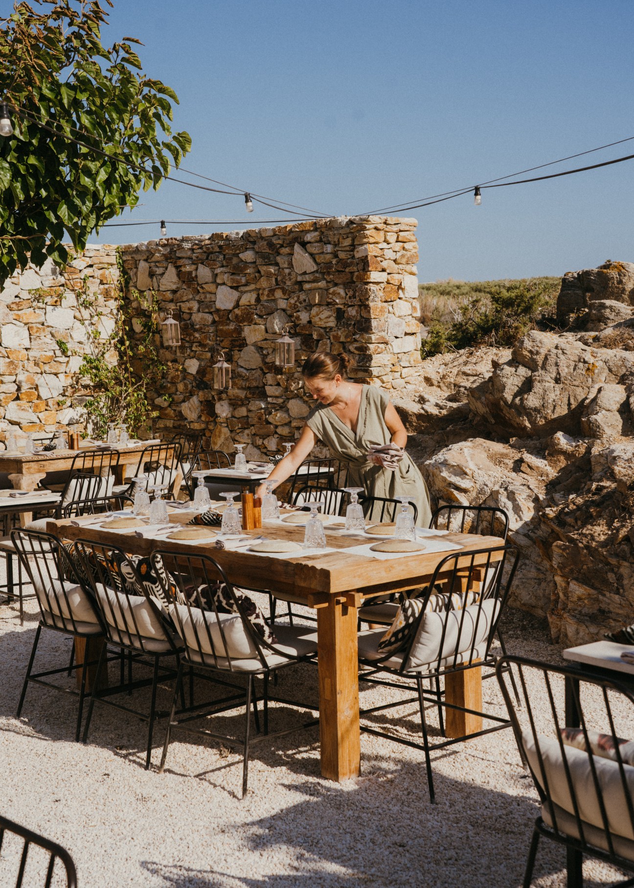 Woman in beige dress setting table at outdoor restaurant terrace with stone walls and string lights