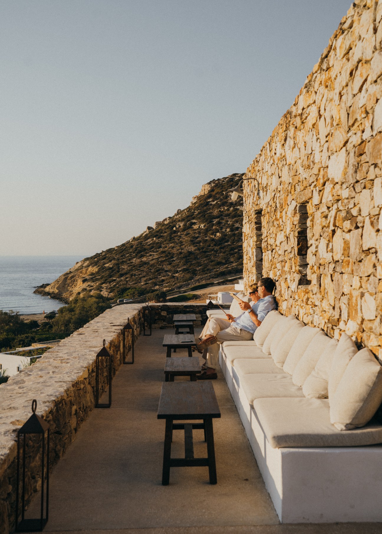 Two guests relaxing on white cushioned seating at The Rooster's bar overlooking the Aegean Sea at sunset
