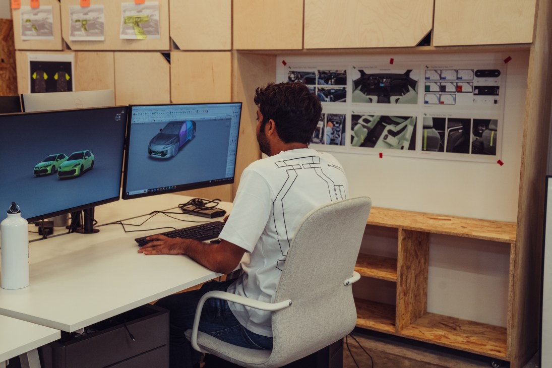 Designer working at desk with dual monitors displaying electric vehicle renderings and design sketches.