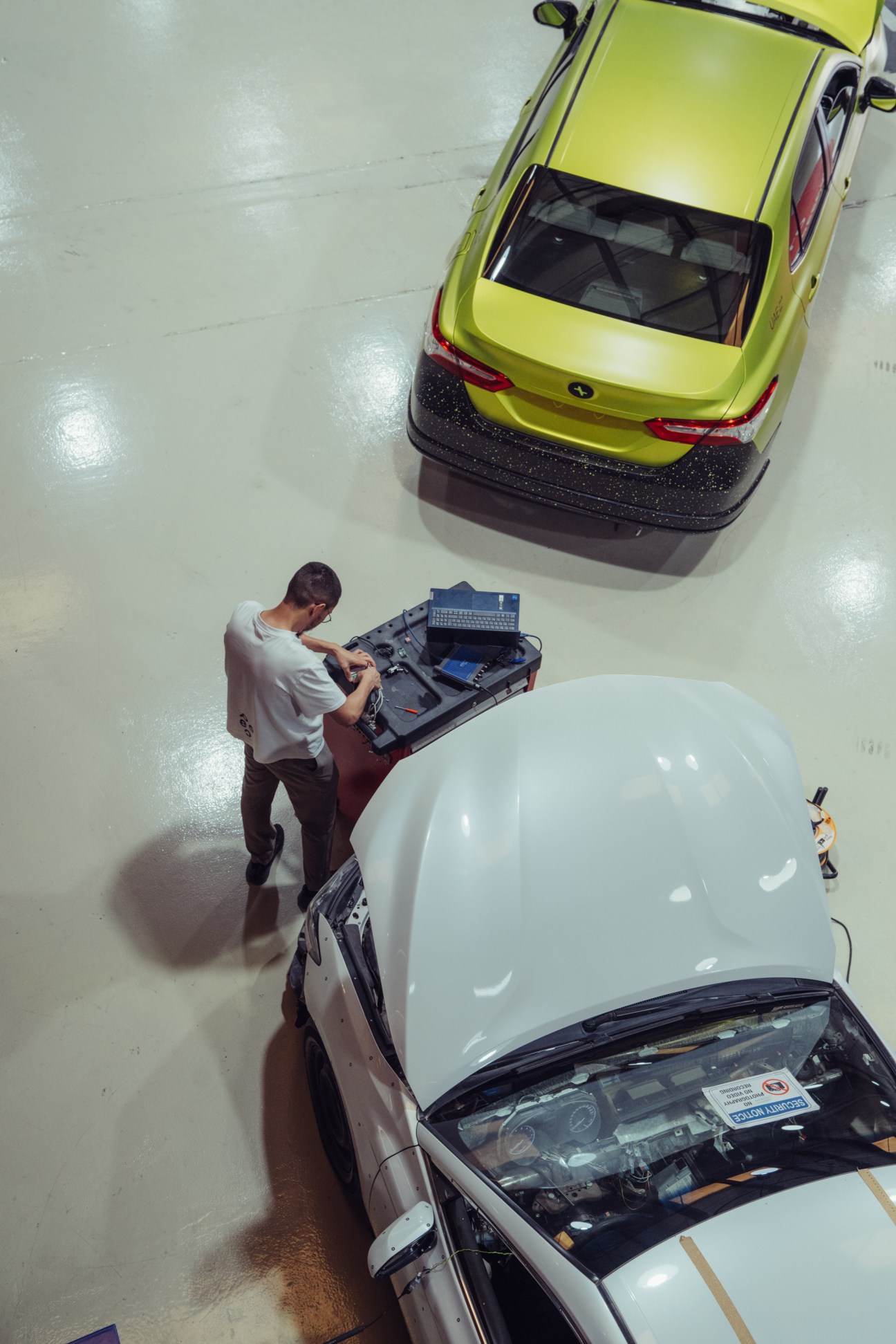 Overhead view of Peec electric vehicle prototypes in a workshop, including a yellow taxi and white sedan with open hood.