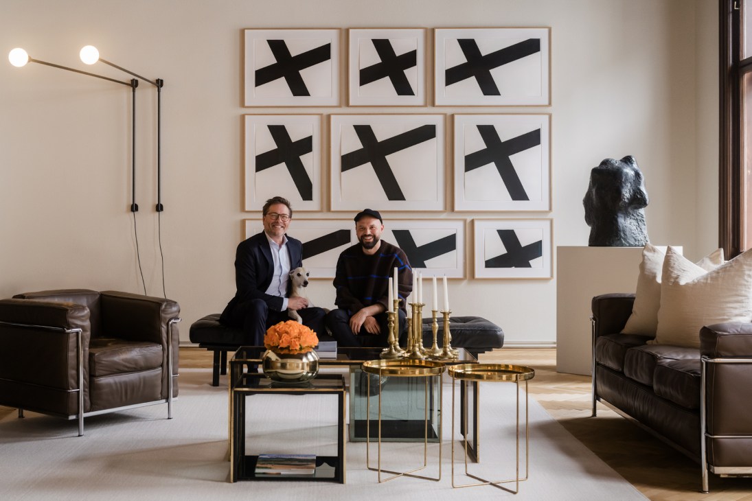 Two men sitting on a leather bench in front of a wall displaying nine framed artworks with black X symbols.