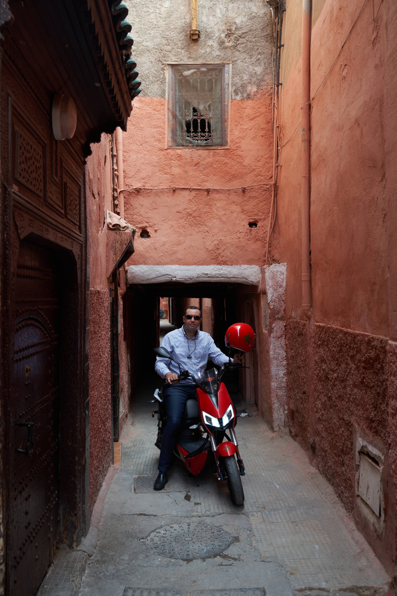 Man in blue shirt and sunglasses riding red electric scooter through narrow alleyway with traditional reddish walls