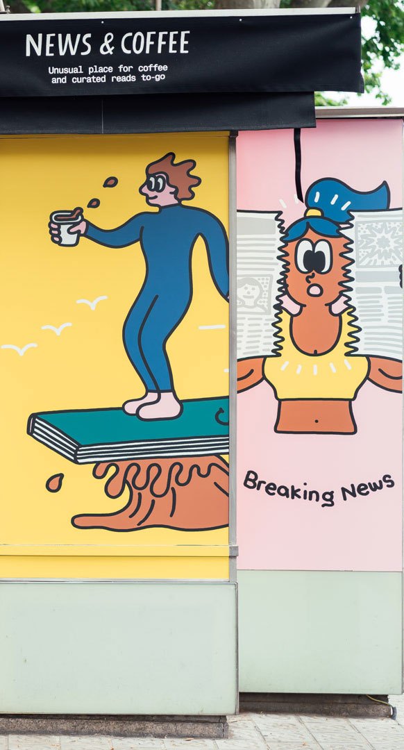 Storefront window display for "News & Coffee" shop featuring colorful illustrated characters: a blue figure holding a coffee cup on a yellow background, and an orange character wearing a blue hat on a pink background labeled "Breaking News