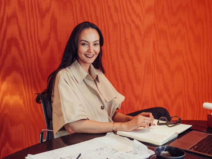 Interior designer Rabah Saeid sits at a desk with architectural drawings and sketches against a vibrant red textured backdrop.
