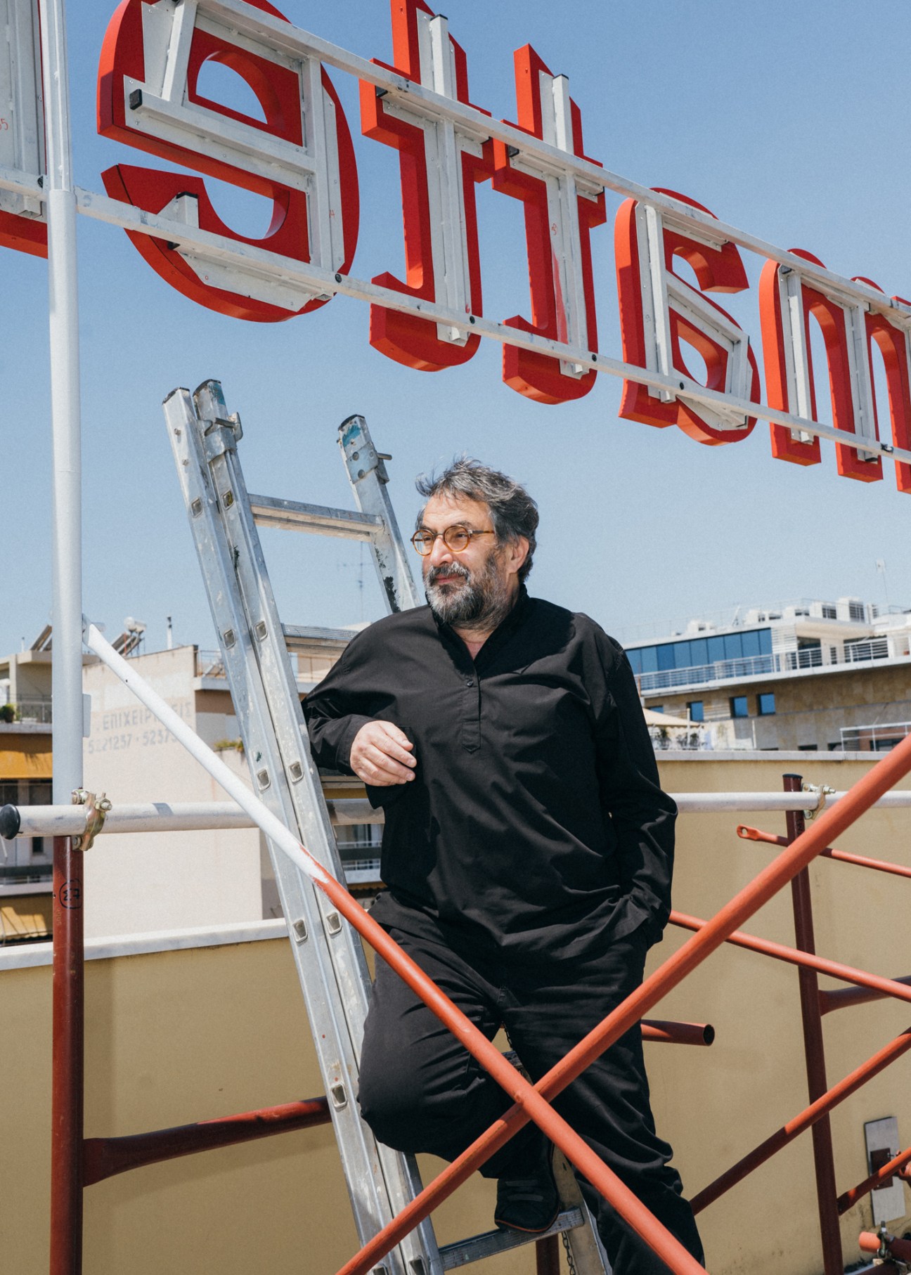 Greek artist Nikos Navridis standing on scaffolding beneath a red neon sign against a blue sky.