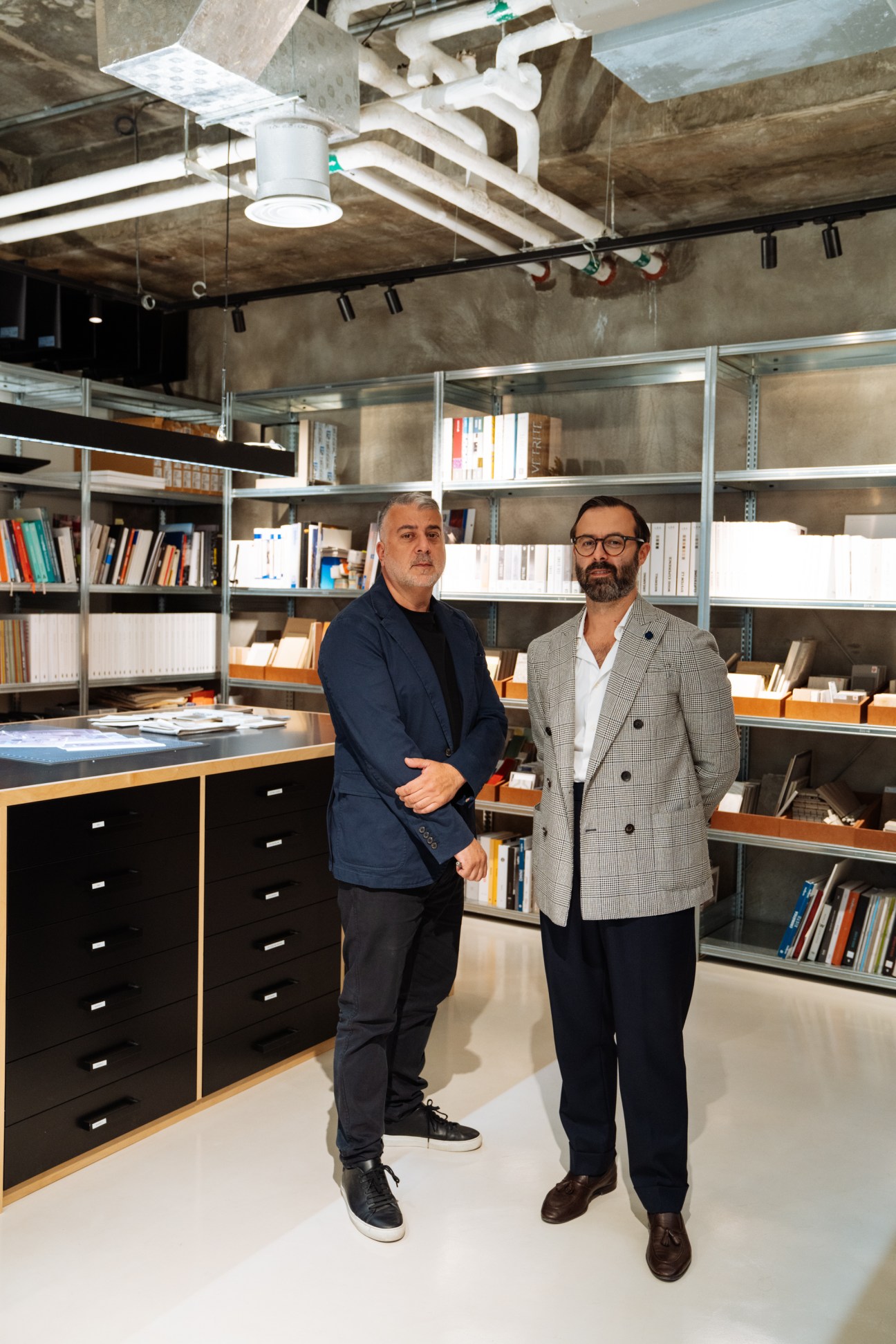 Two men stand in a modern office with industrial ceiling, shelving, and a black desk with drawers in an open-plan workspace.