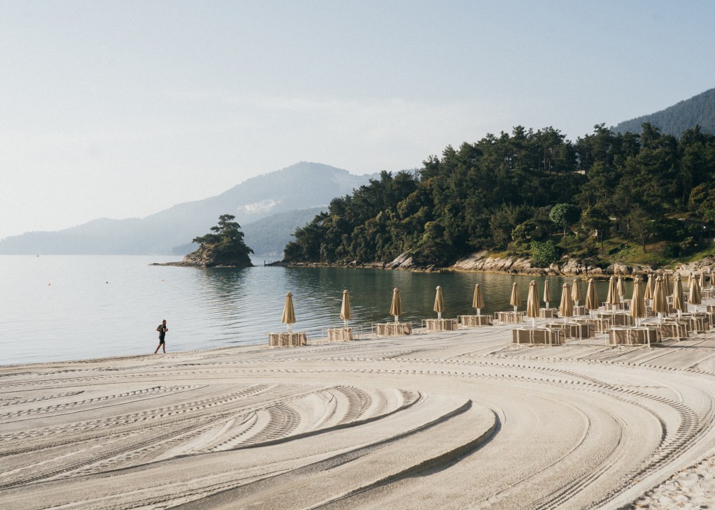 A jogger runs along a pristinely groomed sandy beach with beach umbrellas and chairs arranged in rows, forested mountains visible across the water.
