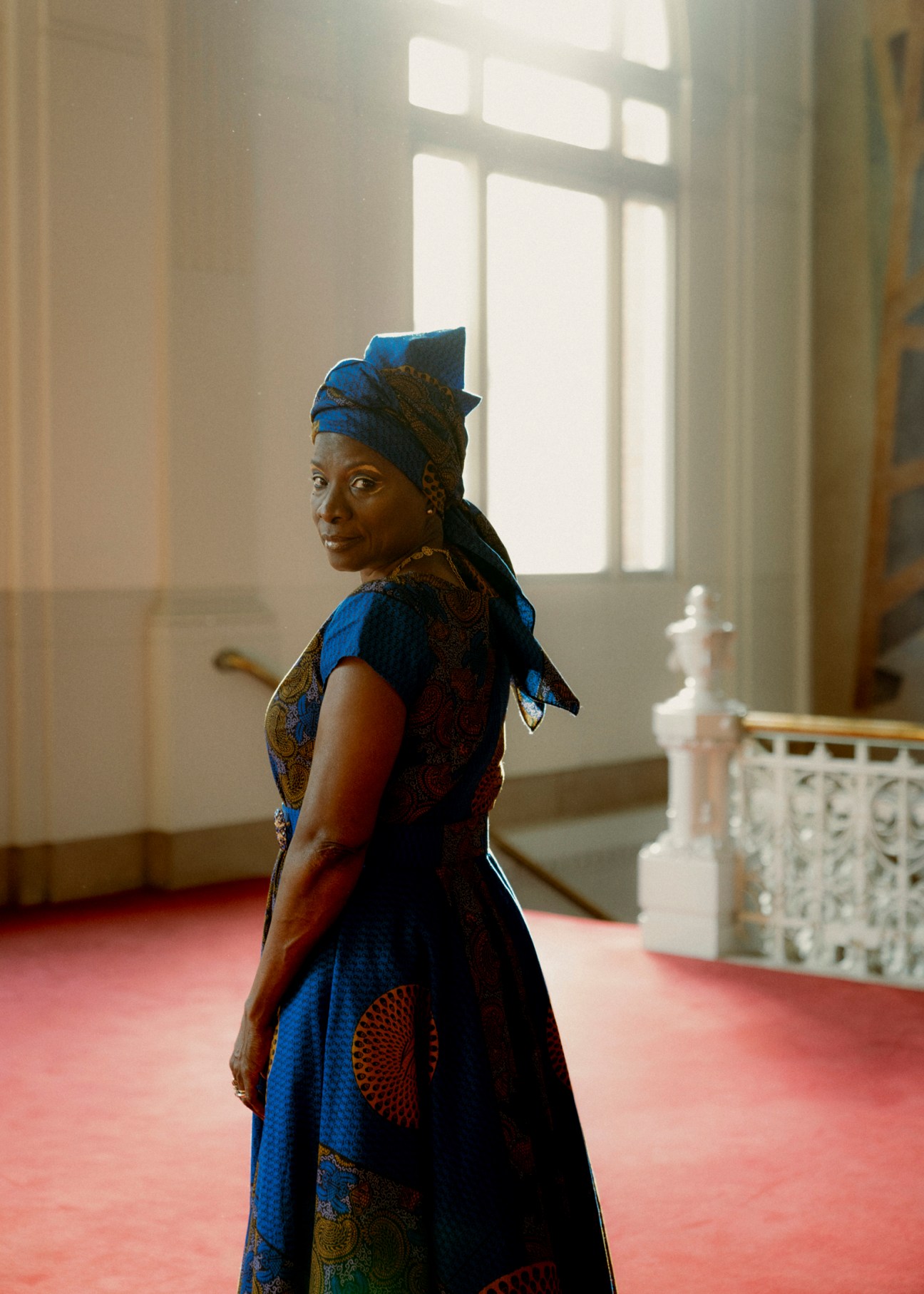 Woman in vibrant blue patterned African dress with matching head wrap standing in elegant interior space