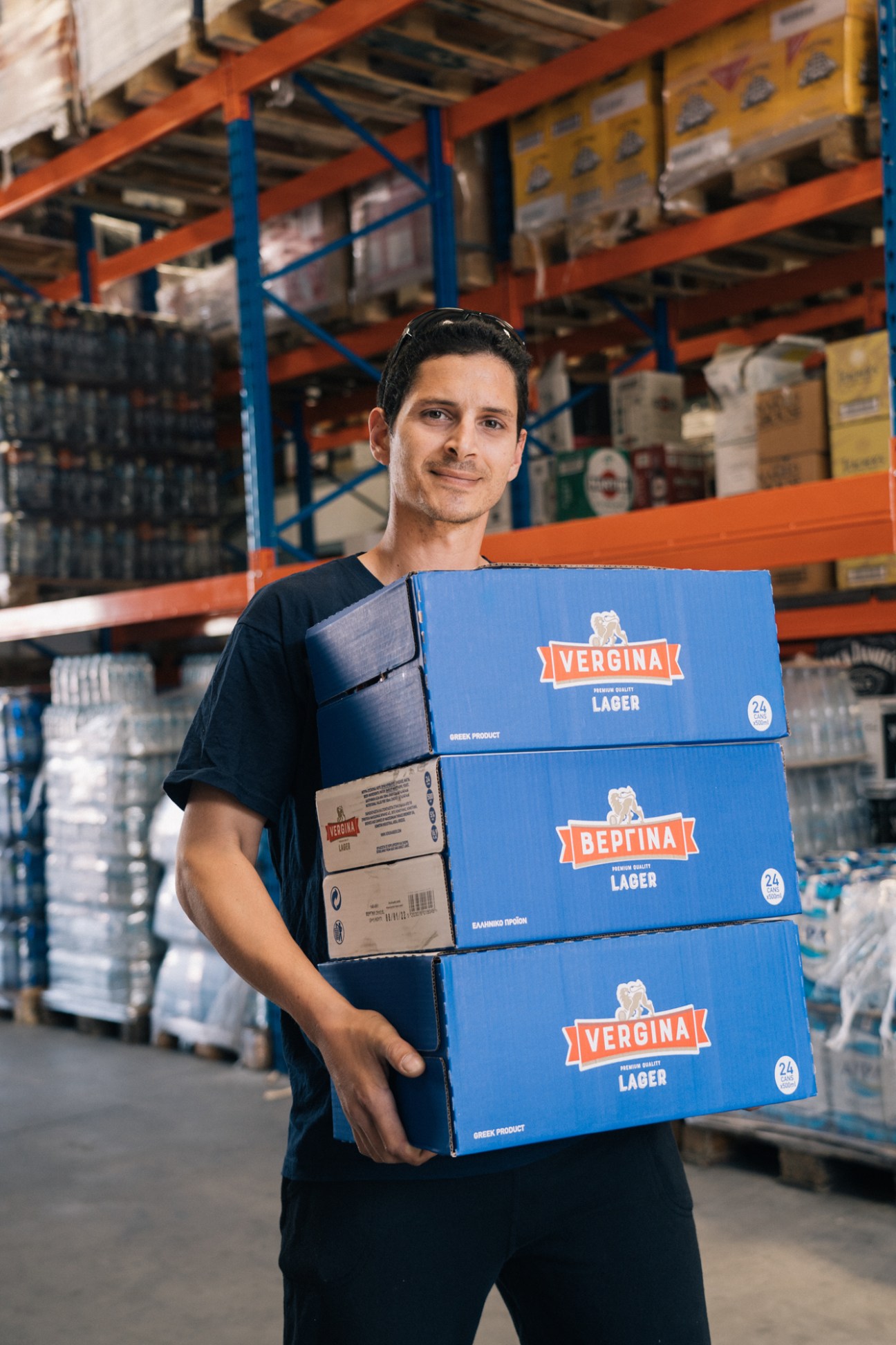 Warehouse worker holding stacked boxes of Vergina lager beer in a distribution depot.