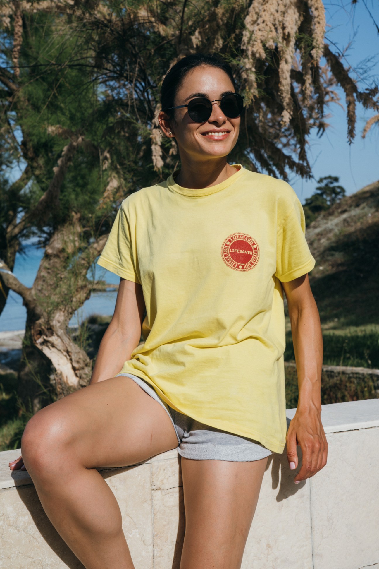 Theodora, a lifeguard at Makryammos hotel, wears a yellow lifesaver t-shirt and sunglasses by a gnarled tree on Thassos beach.