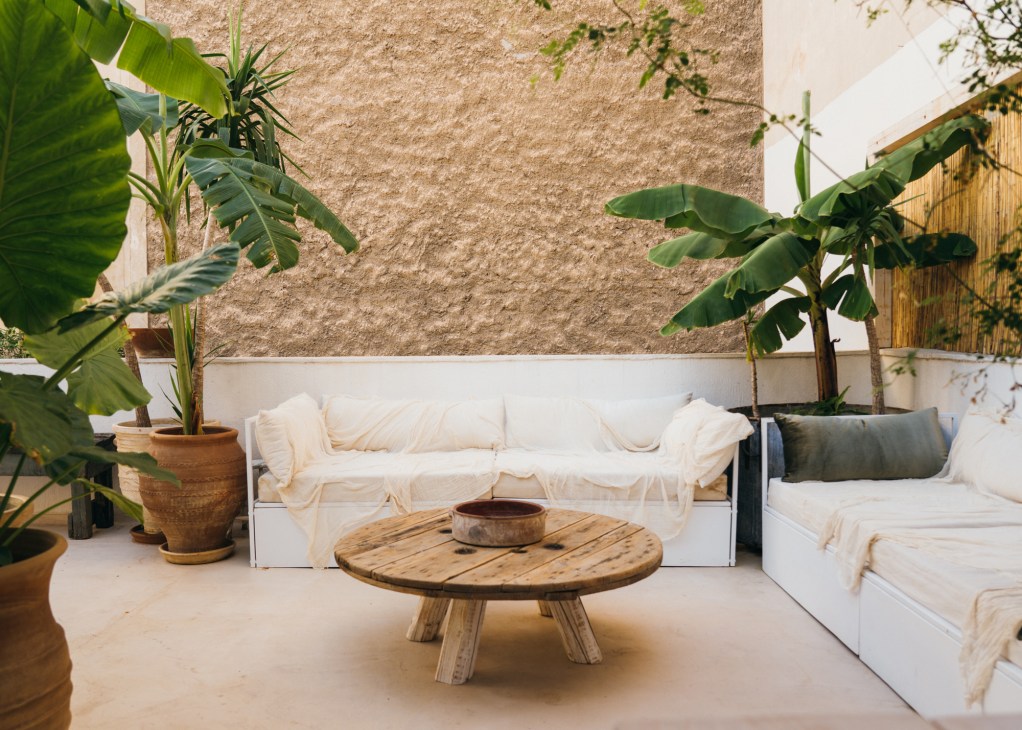 Outdoor terrace seating area with white cushioned benches, wooden coffee table, potted banana plants and bamboo screen