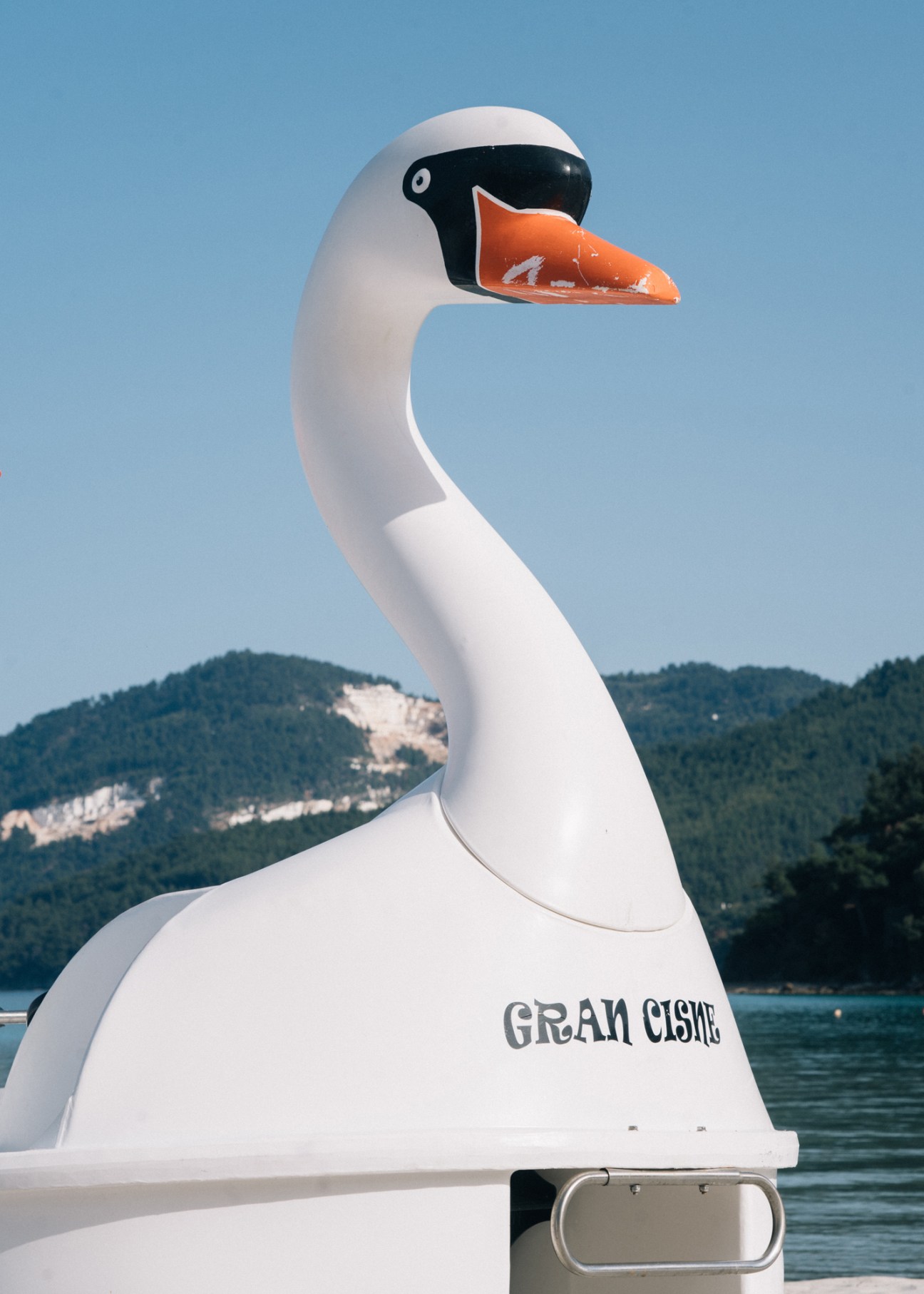 White and orange swan float decorated with 'Gran Cisne' text on a boat with Thassos mountains in the background.