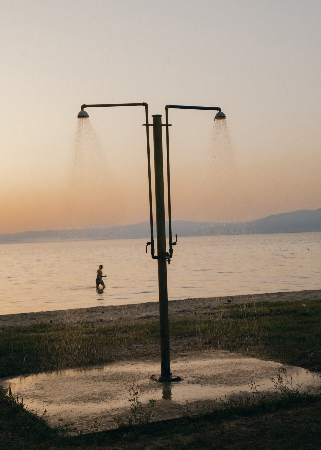 A person wades in the sea at sunset near an outdoor shower on a beach with forested hills beyond.