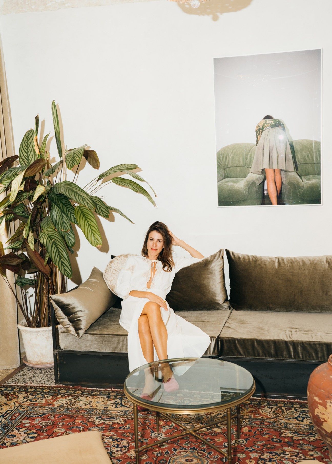 Woman in white dress sitting on olive green velvet sofa in modern living room with large plant and artwork