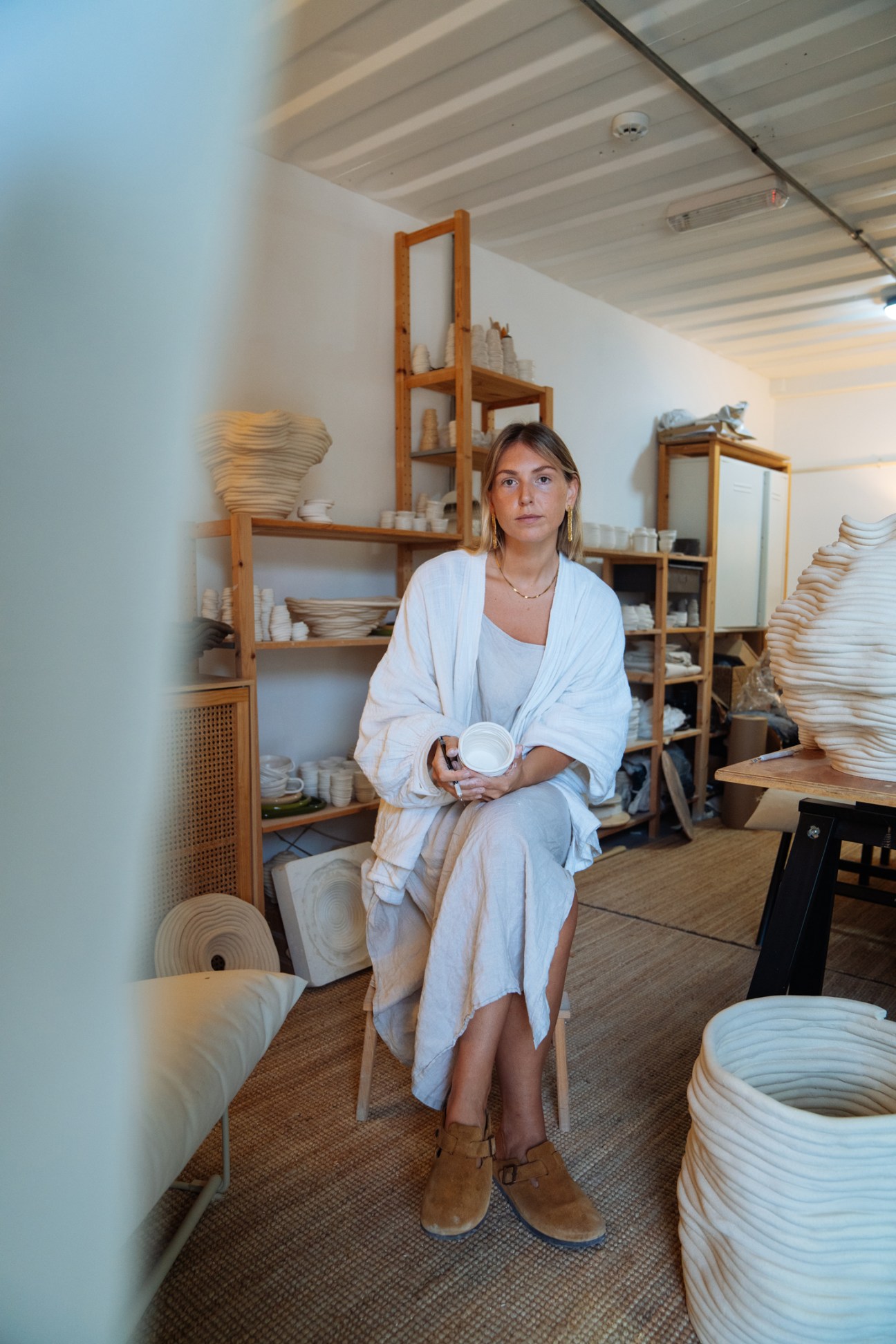Nicole Farrelly sits in her ceramic studio holding a handmade bowl, surrounded by shelves of ceramic pieces.
